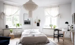 Ett ljust sovrum med en stor taklampa, en dubbelsäng med grå överkast och vita kuddar, samt ett skrivbord med en svart stol.