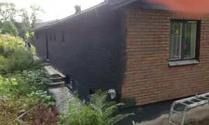 En man håller på att måla tegelväggen svart. En stege står redo vid huset.