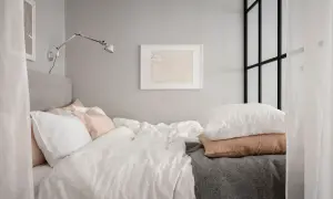 En mysig stuga inredning med en stor säng med vita och rosa kuddar, en grå pläd och en vägglampa.