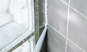 Foga om badrummet med silikon. En fogpistol applicerar ny vit silikon längs en vägg med grå kakelplattor och en duschvägg av glasblock.