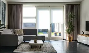 Grå soffa i vardagsrummet med draperade gardiner och stora fönster som släpper in ljus.