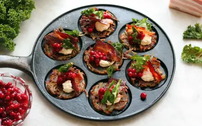 Små, gyllene rårakor i en gjutjärnspanna, toppade med krämig ost, krispigt bacon, lingon och grönkål. Ett läckert råraka recept!