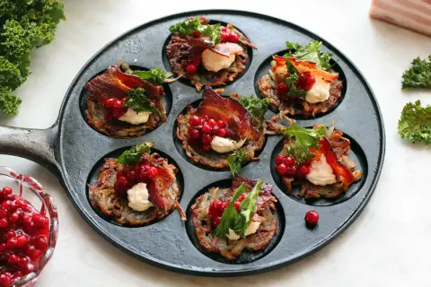 Små, gyllene rårakor i en gjutjärnspanna, toppade med krämig ost, krispigt bacon, lingon och grönkål. Ett läckert råraka recept!