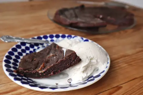 En bit kladdkaka i långpanna serveras med vaniljglass på en blåvit prickig tallrik.