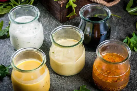 Fem glasburkar med olika dressing till sallad: krämig vit, gul, ljusbrun, mörk och en med frön.