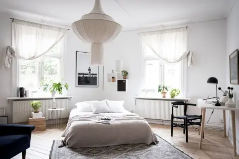 Ett ljust sovrum med en stor taklampa, en dubbelsäng med grå överkast och vita kuddar, samt ett skrivbord med en svart stol.