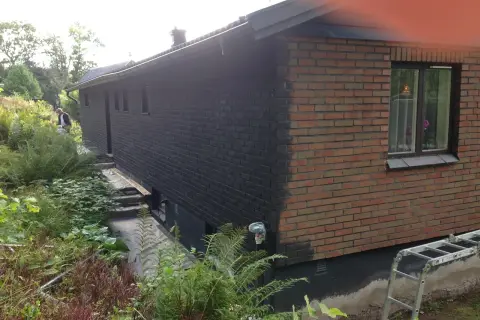 En man håller på att måla tegelväggen svart. En stege står redo vid huset.