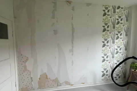 Väggen är delvis avrivet tapet, med fläckar av gammal tapet kvar. En bit med blommig tapet syns till höger.