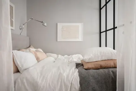 En mysig stuga inredning med en stor säng med vita och rosa kuddar, en grå pläd och en vägglampa.