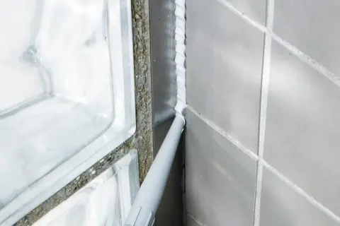 Foga om badrummet med silikon. En fogpistol applicerar ny vit silikon längs en vägg med grå kakelplattor och en duschvägg av glasblock.