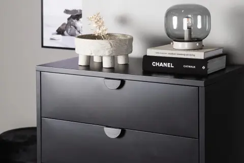 Svart byrå med två lådor, perfekt som byrå sovrum inspiration. Ovanpå står en kruka med torkad växt och en lampa på böcker.