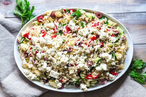 En stor skål med orzo pastasallad, toppad med fetaost, oliver, gurka och paprika. Ett perfekt pastasallad recept för sommaren.