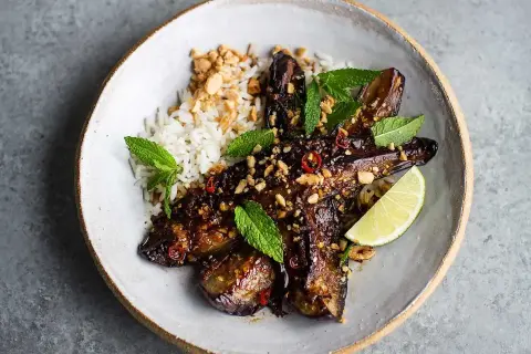 En skål med ris och glaserad, grillad aubergine toppad med jordnötter, chili och mynta. En limeklyfta ligger bredvid.