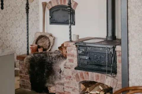 En hund kikar fram bakom en fårskinnspläd vid en gammal gjutjärnskamin. Perfekt lantlig inredning inspiration med tegel och rustika detaljer.