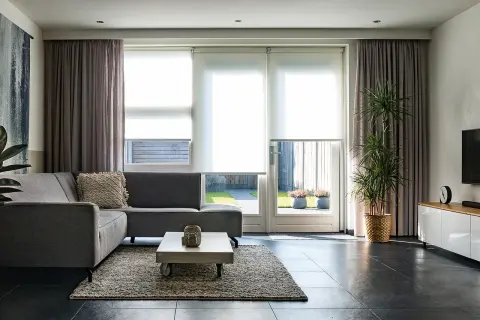 Grå soffa i vardagsrummet med draperade gardiner och stora fönster som släpper in ljus.