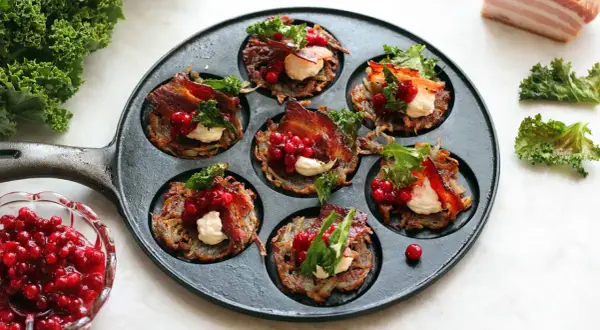 Små, gyllene rårakor i en gjutjärnspanna, toppade med krämig ost, krispigt bacon, lingon och grönkål. Ett läckert råraka recept!