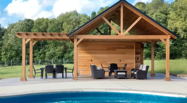 En stor träpergola med tak och sittgrupper vid en blå pool. Kanske är det här man behöver pergola bygglov för att skapa en perfekt uteplats.