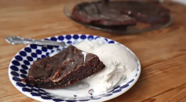 En bit kladdkaka i långpanna serveras med vaniljglass på en blåvit prickig tallrik.