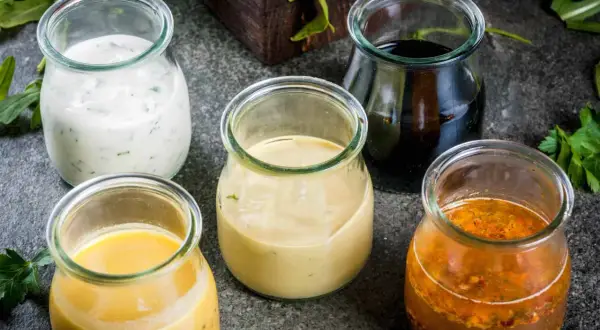 Fem glasburkar med olika dressing till sallad: krämig vit, gul, ljusbrun, mörk och en med frön.