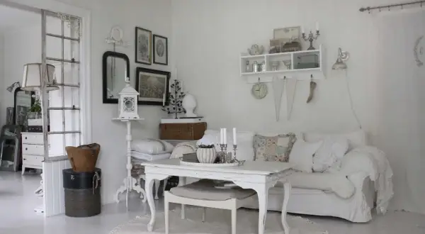 Ett rum med shabby chic inredning. En vit soffa med kuddar, ett vitt bord med ljus och en rund matta.