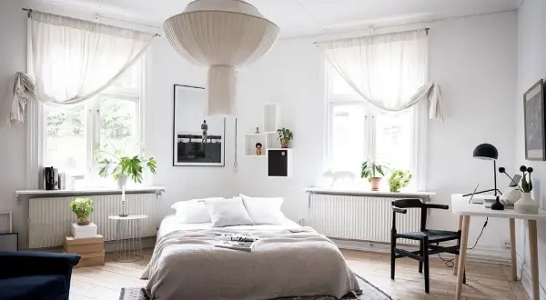 Ett ljust sovrum med en stor taklampa, en dubbelsäng med grå överkast och vita kuddar, samt ett skrivbord med en svart stol.