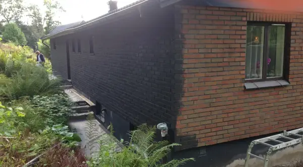 En man håller på att måla tegelväggen svart. En stege står redo vid huset.