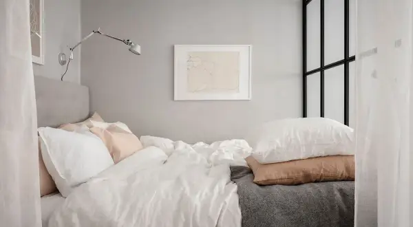 En mysig stuga inredning med en stor säng med vita och rosa kuddar, en grå pläd och en vägglampa.