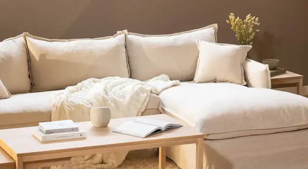 En bekväm soffa med pastell inredning, prydd med mjuka kuddar och en fluffig pläd. Ett soffbord i trä med böcker och en vas fulländar den lugna atmosfären.