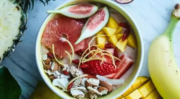 En skål med nyttiga smoothies fylld med jordgubbar, mango, grapefrukt, nötter och kokosflingor. Omgiven av färsk frukt som ananas och banan.