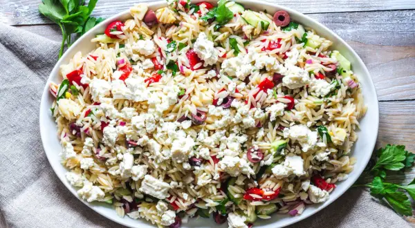 En stor skål med orzo pastasallad, toppad med fetaost, oliver, gurka och paprika. Ett perfekt pastasallad recept för sommaren.