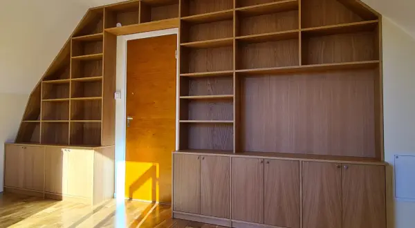 En stor, skräddarsydd bokhylla i trä som byggts in i ett snedtak. Den har många hyllplan och skåp, perfekt för att bygga en ny förvaringslösning.