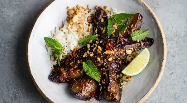 En skål med ris och glaserad, grillad aubergine toppad med jordnötter, chili och mynta. En limeklyfta ligger bredvid.