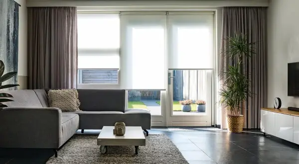Grå soffa i vardagsrummet med draperade gardiner och stora fönster som släpper in ljus.