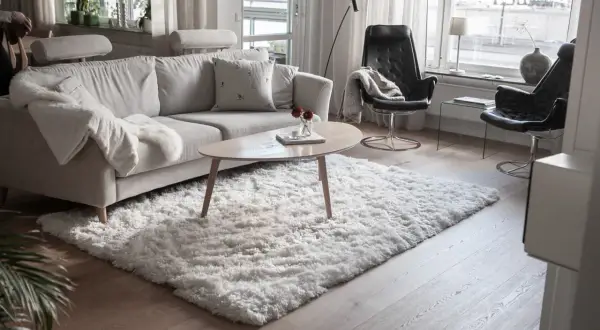 En fluffig, vit matta ligger framför en ljusgrå soffa och skapar en mysig känsla i vardagsrummet.