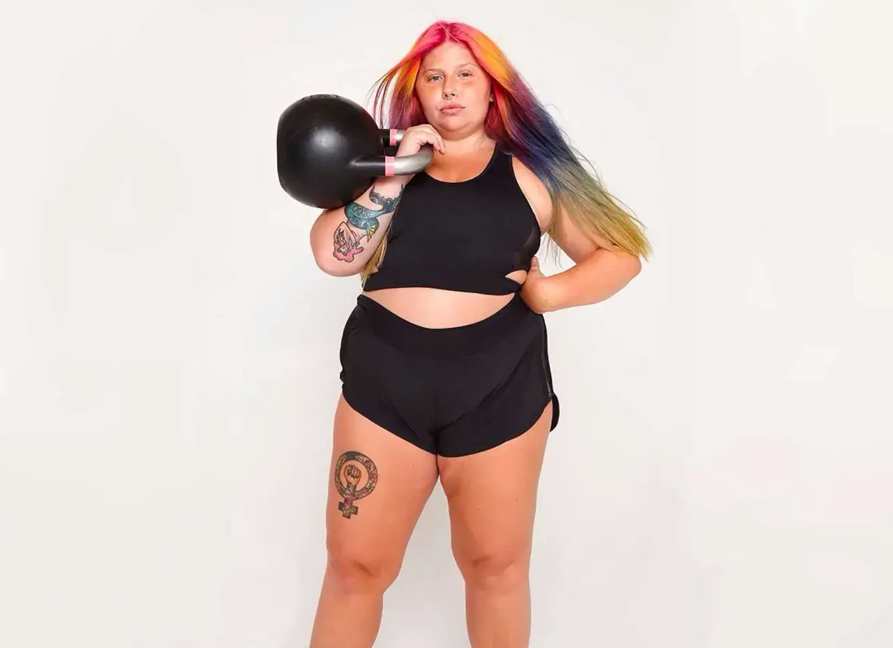 Kvinna med färgglatt hår tränar med kettlebell. Frågan "hur får man smalare lår" kan besvaras med styrketräning som denna.