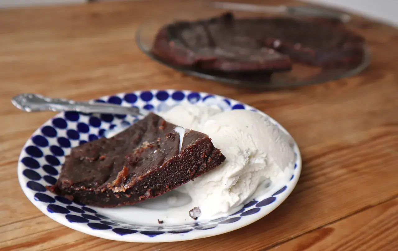 En bit kladdkaka i långpanna serveras med vaniljglass på en blåvit prickig tallrik.