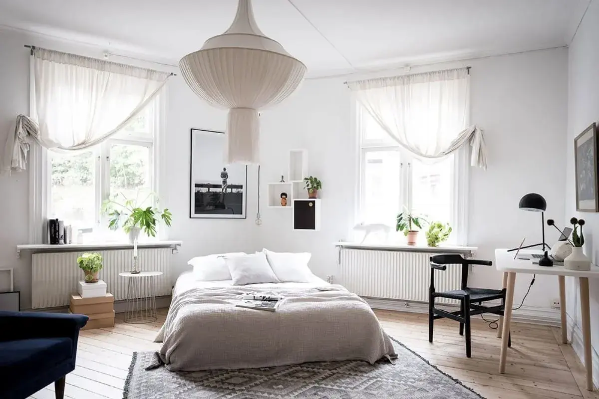 Ett ljust sovrum med en stor taklampa, en dubbelsäng med grå överkast och vita kuddar, samt ett skrivbord med en svart stol.