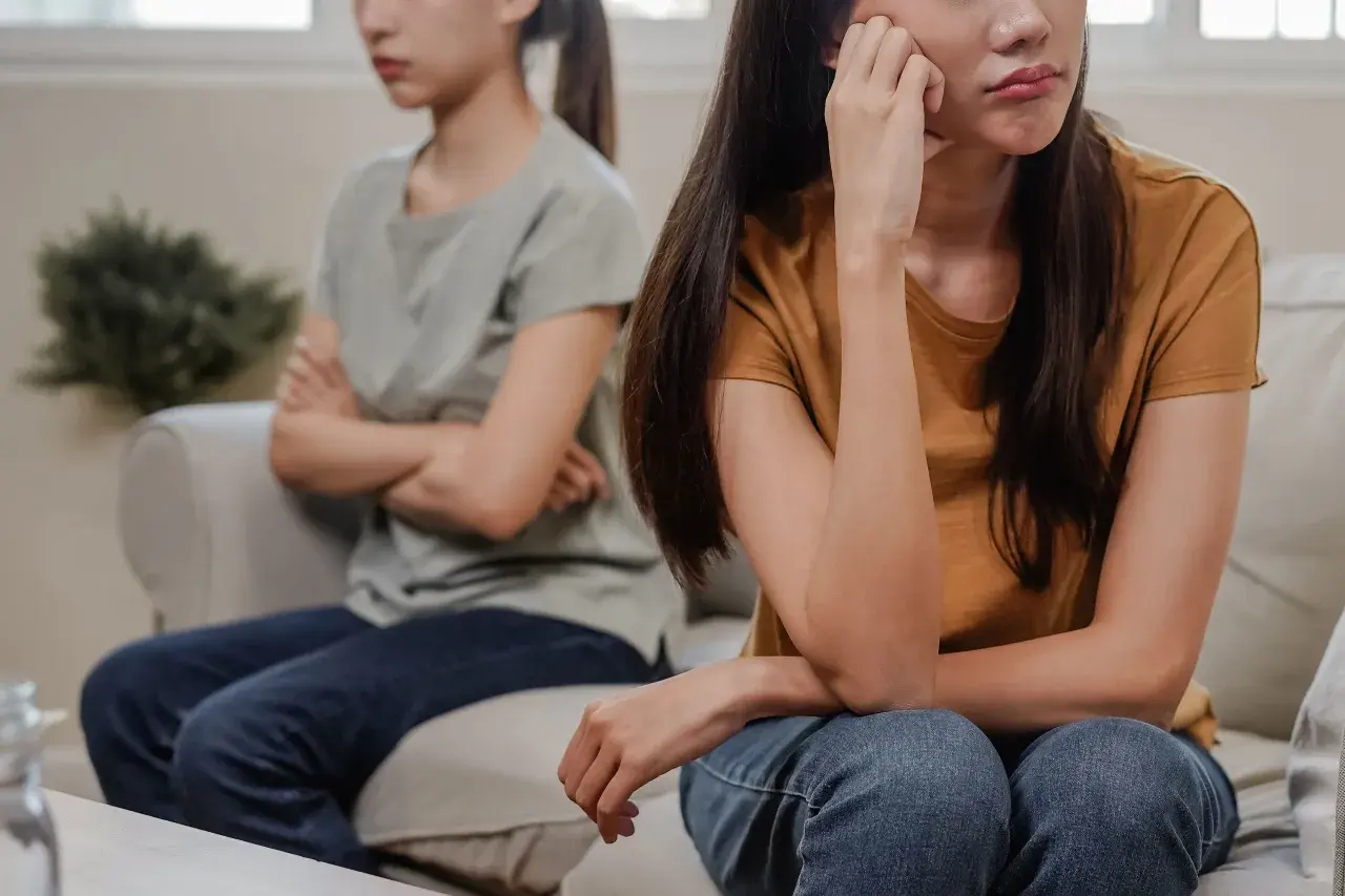 Två systrar sitter på en soffa, vända bort från varandra. Den ena har armarna i kors, den andra vilar hakan i handen. De verkar vara syskon som inte vill umgås.