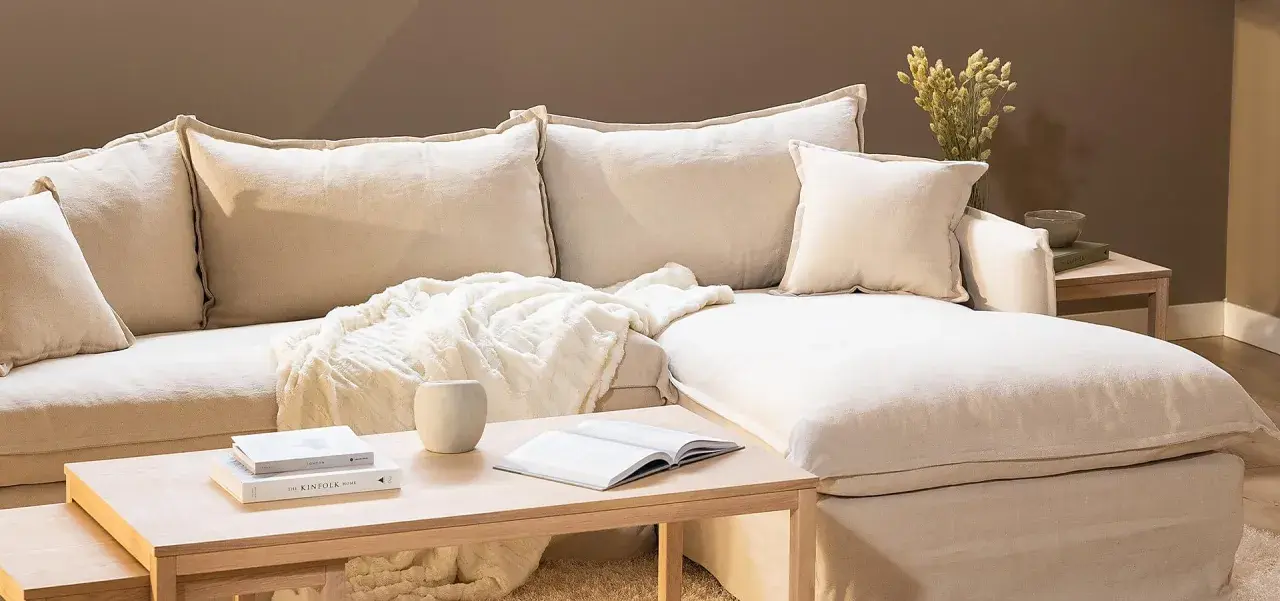En bekväm soffa med pastell inredning, prydd med mjuka kuddar och en fluffig pläd. Ett soffbord i trä med böcker och en vas fulländar den lugna atmosfären.
