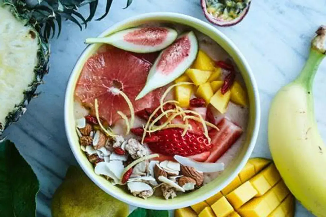 En skål med nyttiga smoothies fylld med jordgubbar, mango, grapefrukt, nötter och kokosflingor. Omgiven av färsk frukt som ananas och banan.