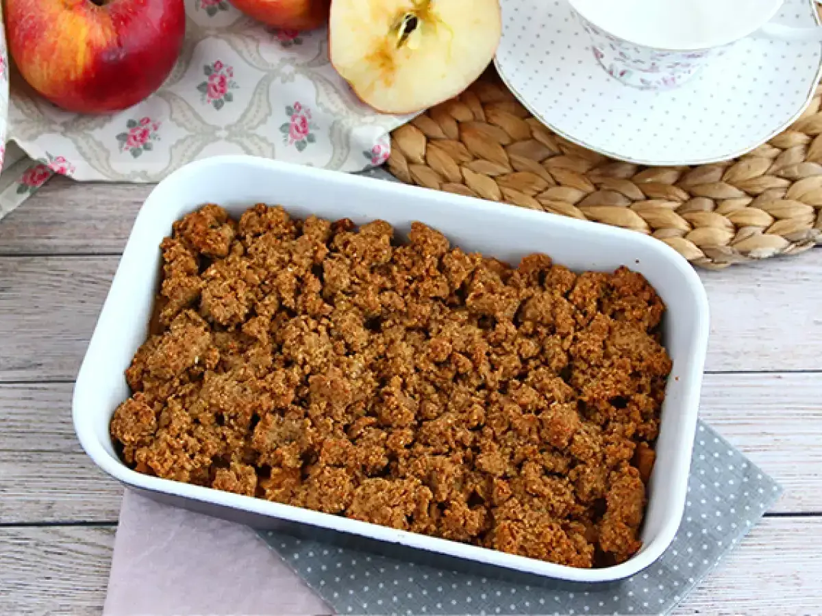 En härlig glutenfri frukost med äppelcrisp i en vit form, äpplen och en kopp te i bakgrunden.