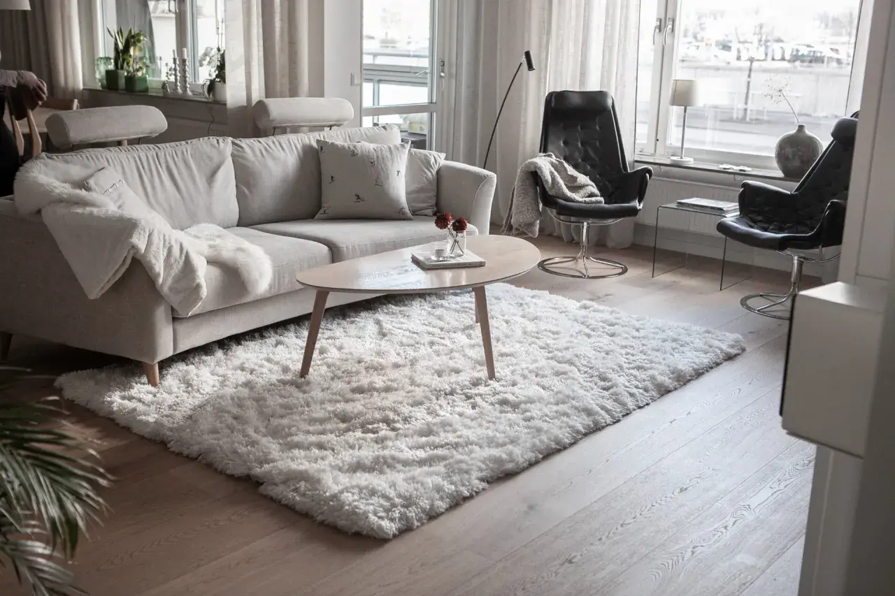 En fluffig, vit matta ligger framför en ljusgrå soffa och skapar en mysig känsla i vardagsrummet.