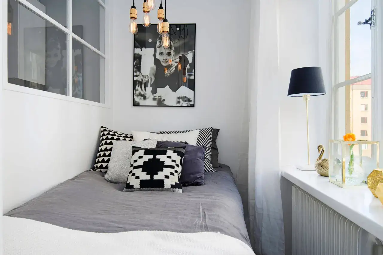 Snyggt litet sovrum inspiration med Audrey Hepburn-poster, grå sängkläder och grafiska kuddar. En guldfärgad svan och en blomma pryder fönsterbrädan.