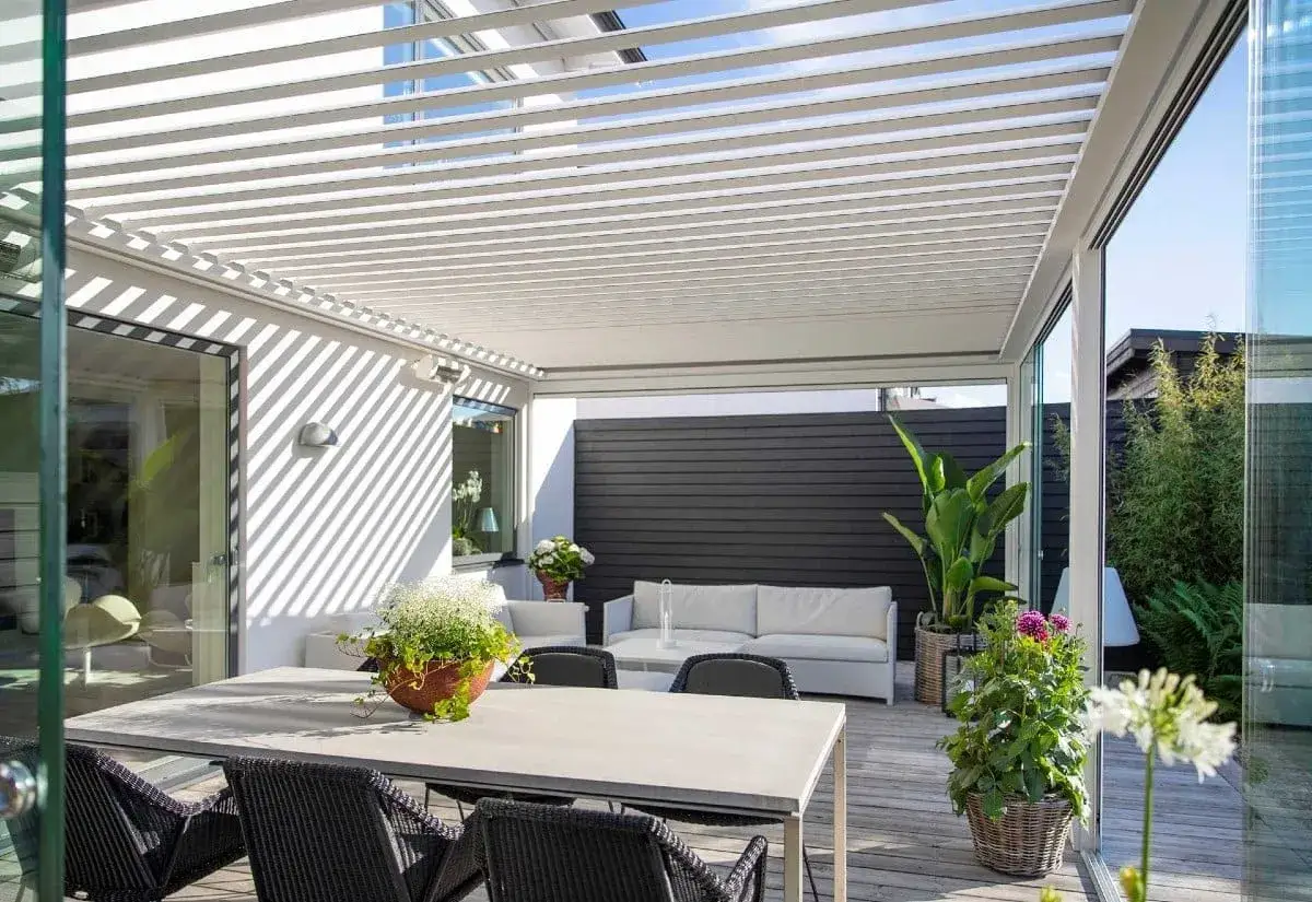 En modern uteplats med ett stilrent tak f&ouml;r att bygga pergola med tak. H&auml;r finns matbord, soffgrupp och gr&ouml;na v&auml;xter.