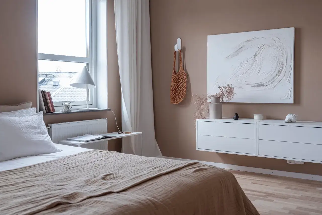 Ett ljust sovrum med en stor säng med beige överkast, en vit lampa vid fönstret och en vit vägghylla med konst och dekorationer.