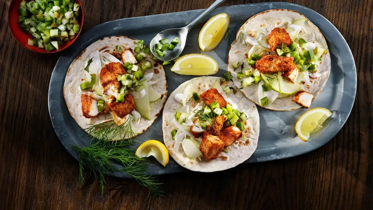 Tre l&auml;ckra fish tacos med krispig fisk, gr&ouml;nsaker och citronklyftor p&aring; ett fat.