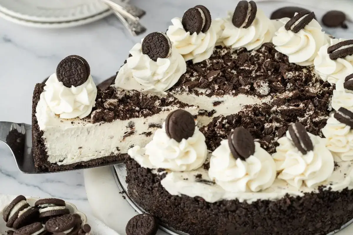 En kr&auml;mig oreo cheesecake med vispgr&auml;dde och hela oreos. En bit lyfts upp med en t&aring;rtspade.