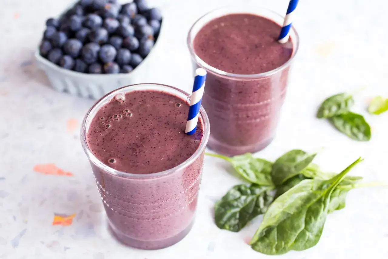 Tv&aring; glas med lila, nyttiga smoothies. En sk&aring;l med bl&aring;b&auml;r och n&aring;gra spenatblad ligger bredvid.