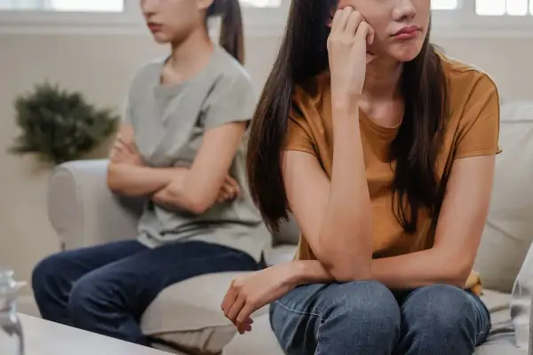 Två systrar sitter på en soffa, vända bort från varandra. Den ena har armarna i kors, den andra vilar hakan i handen. De verkar vara syskon som inte vill umgås.