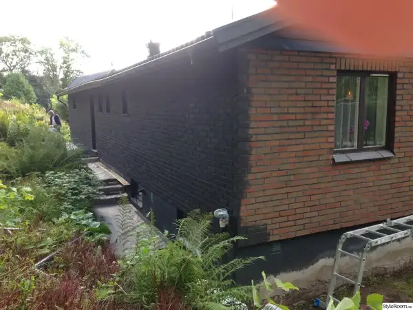 En man håller på att måla tegelväggen svart. En stege står redo vid huset.